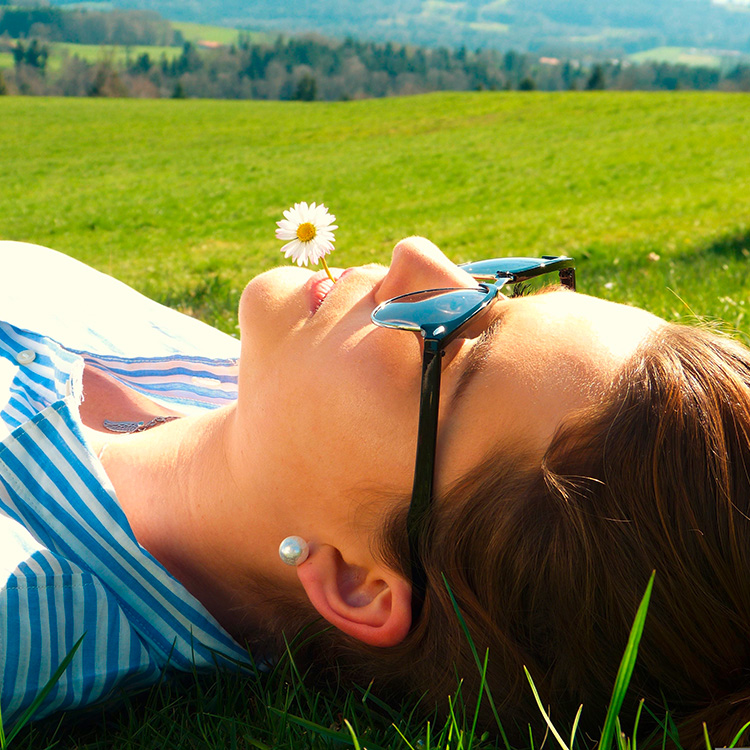 Junge Frau liegt in der Sonne auf einer Wiese mit einem Gänseblümchen zwischen den Lippen.